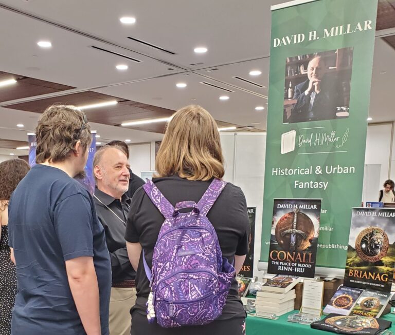 Author David H. Millar engages with fans at a book fair, showcasing his historical and urban fantasy works displayed on a table.