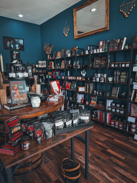 A cozy shop interior features dark blue walls filled with shelves of books, candles, and eclectic decor. A wooden table displays unique items, inviting exploration.