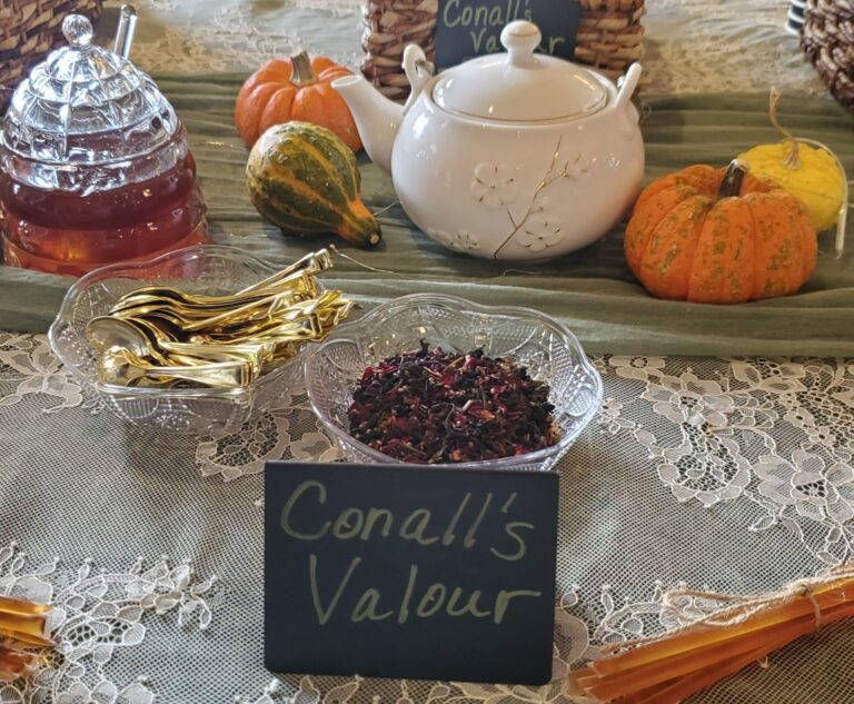 A teapot and honey jar with golden spoons and a dish of dried flowers labeled "Conall’s Valour," surrounded by small pumpkins on a lace tablecloth.