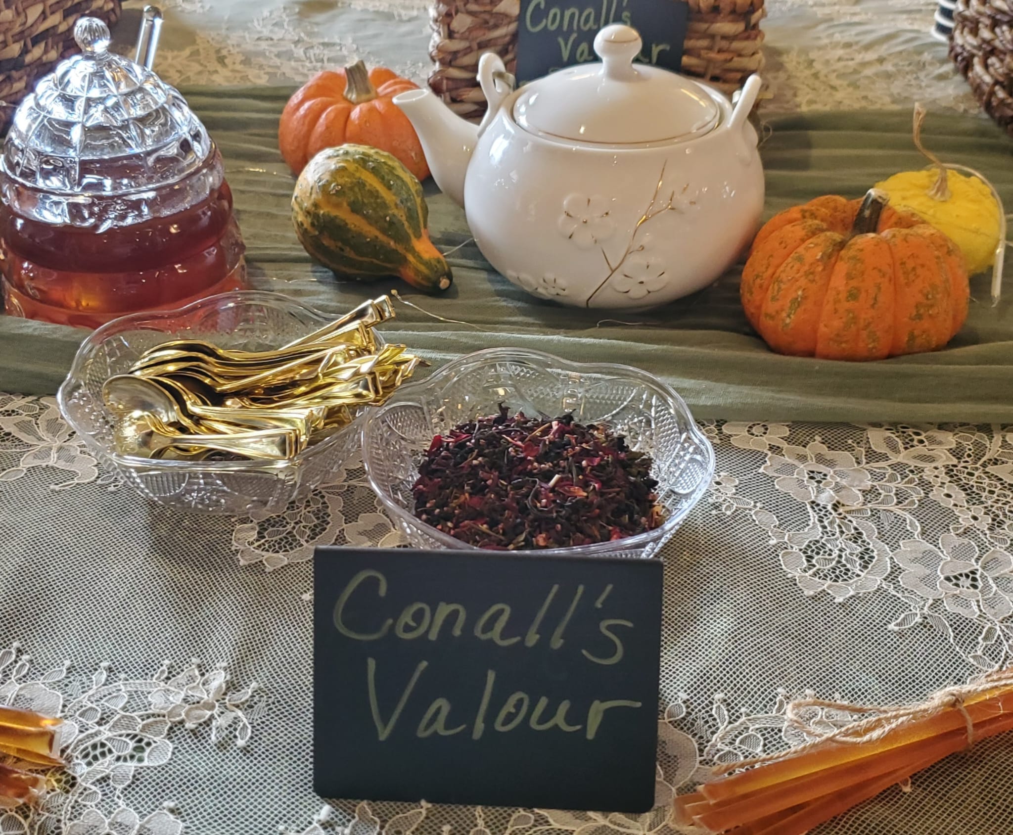 A teapot and honey jar with golden spoons and a dish of dried flowers labeled "Conall’s Valour," surrounded by small pumpkins on a lace tablecloth.